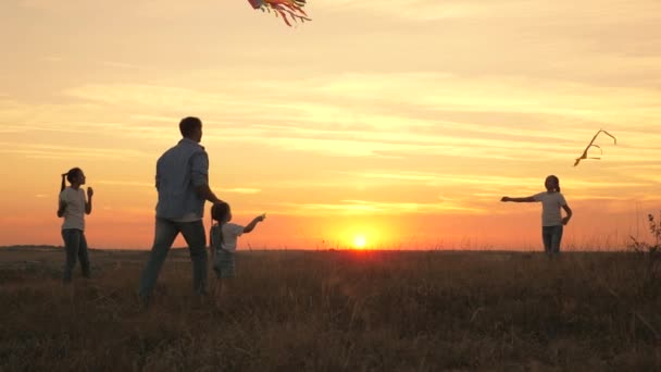 Tata z dziećmi bawi się latawcami o zachodzie słońca w parku. Rodzinna gra na świeżym powietrzu. Tatuś i zdrowe córki wystrzeliwują w niebo kolorowe żywice papierowe. zabawa z rodzicami w słońcu — Wideo stockowe