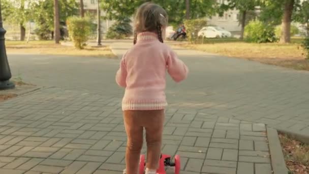 Een klein meisje leert op een scooter rijden. Een gelukkig meisje dat in het park speelt. gezond kind rijdt op een scooter in de stad op straat. gelukkige kindertijd concept — Stockvideo