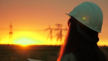 Beyaz kasklı bir kadın güç mühendisi tabletteki elektrik sensörlerinden elde edilen verileri kullanarak güç hattını kontrol ediyor. Gün batımında yüksek voltajlı elektrik hatları. Dağıtım ve elektrik tedariği. temiz enerji