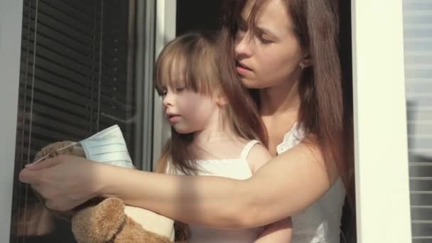 Familie kind en moeder in quarantaine. Mam en dochter spelen op vensterbank, kleden teddybeer met een beschermend masker tegen virussen, kijken uit het raam. Een kind knuffelt een speeltje. Covid-19 — Stockvideo