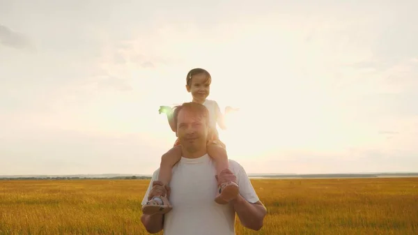 Papa draagt op zijn schouders zijn geliefde kleine gezonde dochter in de zon. Vader loopt met het kind op zijn schouders bij zonsondergang. kind met ouders die in het veld lopen. gelukkig gezin rust uit in park. — Stockfoto