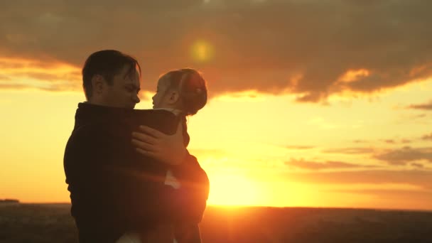 Szczęśliwe dziecko i ojciec bawią się na polu w słońcu. Tata przytula i przykrywa swoją córeczkę kurtką w chłodny wieczór w parku o zachodzie słońca. zdrowe dziecko całuje tatusia. szczęśliwa rodzina i koncepcja dzieciństwa — Wideo stockowe