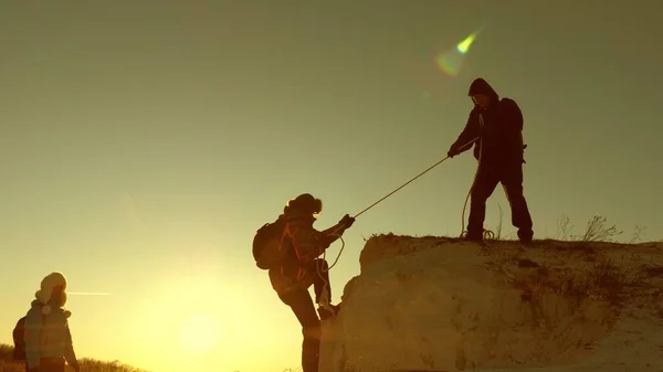 Scalatori scalano la montagna su una corda. un turista maschio aiuta una ragazza viaggiatore a scalare la montagna. Viaggio e avventura in montagna al tramonto. lavoro di squadra di turisti al sole. concetto di libertà e avventura — Foto Stock