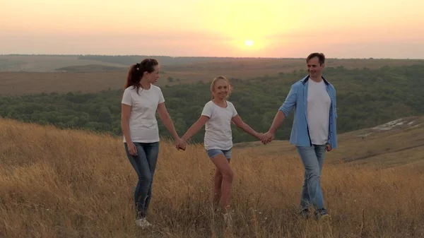 Здоровая улыбающаяся семья, держащаяся за руки, гуляющая в поле на закате в горах, дочь, держащая маму и папу за руки. счастливый ребенок и родители ходят в лучах красивого солнца, путешествовать в отпуск — стоковое фото