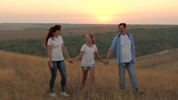 Zdrowa uśmiechnięta rodzina trzymająca się za ręce spacerujące po polu o zachodzie słońca w górach, córka trzymająca mamę i tatę za ręce. szczęśliwe dziecko i rodzice chodzić w promieniach pięknego słońca, podróżować na wakacje — Zdjęcie stockowe