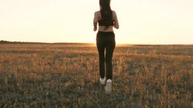 Sağlıklı, güzel bir kız spor yapıyor, kırsalda güneş altında koşuyor. Antrenman koşusu. Koşucu kız sahada temiz hava soluyor. Özgür genç kadın gün batımında yaz parkında koşuyor..