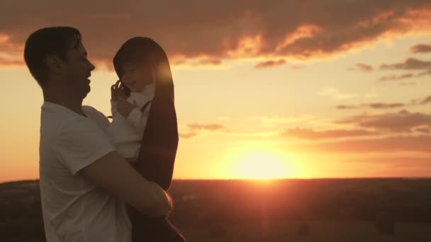 Papa umarmt und bedeckt seine kleine Tochter an einem kühlen Abend im Park bei Sonnenuntergang mit Jacke. gesundes Baby küsst Papa. Glückliches Kind und Vater spielen in der Sonne auf dem Feld. glückliche Familie und Kindheit — Stockvideo