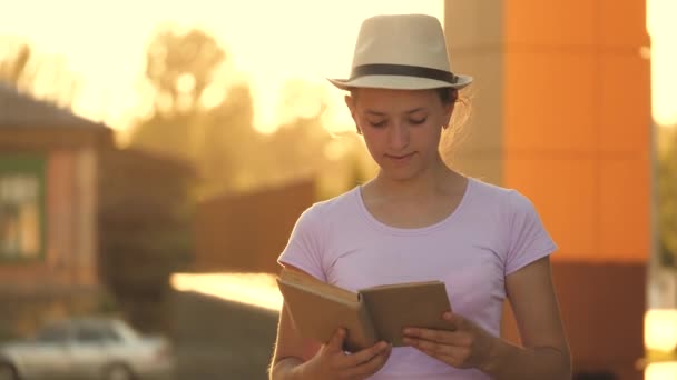 Een tienermeisje met een hoed loopt door de straat in de zon en leest een boek, in de stad in de buurt van het schoolgebouw — Stockvideo