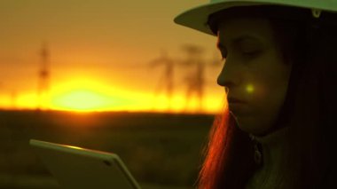 Gün batımında yüksek voltajlı elektrik hatları. Temiz enerji. Beyaz kasklı bir kadın güç mühendisi tabletteki elektrik sensörlerinden elde edilen verileri kullanarak güç hattını kontrol ediyor..