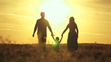 Mutlu çocuk ailesi kavramı. Sağlıklı küçük kız gün batımında bir tarlada anne ve babasının kollarında atlayıp uçuyor. Mutlu aile güneşin altında parkta yürür. Anne, baba ve bebek dışarıda yürüyor..