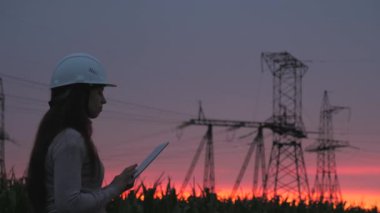 Dağıtım ve elektrik tedariği. Temiz enerji. Beyaz kasklı bir kadın güç mühendisi tabletteki elektrik sensörlerinden elde edilen verileri kullanarak güç hattını kontrol ediyor. Gün batımında yüksek voltajlı elektrik hatları