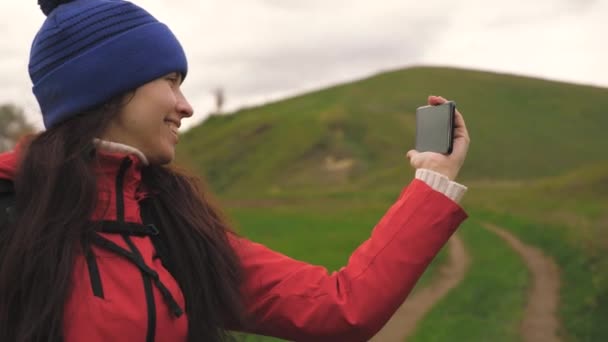 Junge Touristenbloggerin filmt Selfie-Videos am Fuße der Berge mit einem Smartphone mit einer wunderschönen Landschaft im Hintergrund. Gesunde, fröhliche Frau reist und fotografiert die Natur per Telefon. — Stockvideo