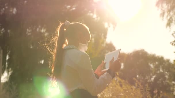 Meisje tiener loopt met een tablet in de straat in het park in de stralen van de warme zon. Portret van een jong meisje met een tablet in de hand in een straat. Gelukkig kind online op het internet — Stockvideo