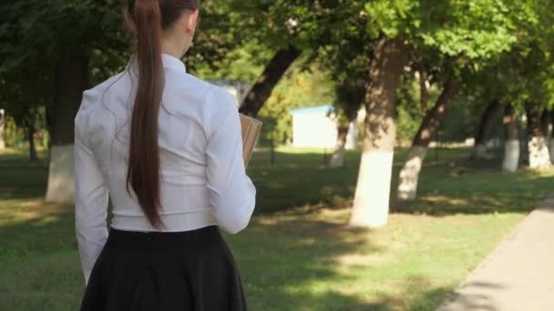 Adolescente menina apressa-se para a escola com livros. Uma estudante caminha pela rua com livros didáticos nas mãos no parque de verão. estudante na cidade. — Vídeo de Stock