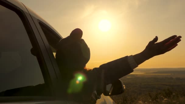 O fată sănătoasă își flutură mâna de la mașină și admiră apusul de soare frumos de pe munte. Călători liberi, turiști. femeie călător călătorește cu mașina admirând răsăritul soarelui, pădurea și râul de la fereastra mașinii . — Videoclip de stoc