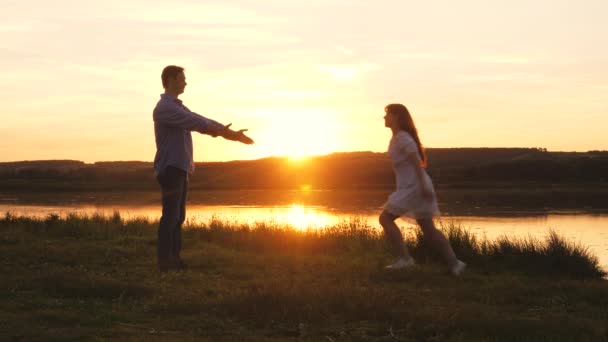 Boldog lány beleugrik szeretett pasija karjaiba. Szerelmes férfi és nő táncolnak a nap ragyogó sugaraiban a tó hátterében. A család napnyugtakor táncol a parton. szabad emberek — Stock videók