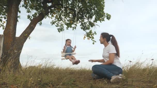 La familia libre se está divirtiendo en el parque. madre sacude a su pequeña hija sana en un columpio bajo un árbol en el sol. madre juega con su hijo, se balancean en la cuerda en una rama de roble en el bosque. — Vídeos de Stock