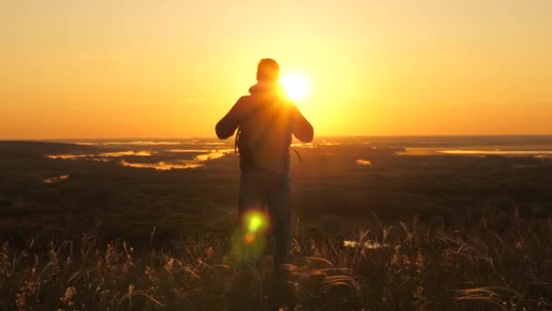 夜明けの光線の山の端にバックパック付きの無料の男性旅行者は、彼の手を上げ、勝利、美しい太陽と風景を楽しんでいます。一人の観光客が自然の中を。冒険 — ストック動画