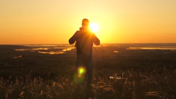 Um viajante masculino livre com uma mochila fica à beira de uma montanha em raios de madrugada, levanta as mãos e desfruta de vitória, o belo sol e paisagem. turista solitário viaja na natureza. Aventuras — Vídeo de Stock