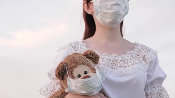 Młoda kobieta nosi ulubioną zabawkę swojego dziecka. pluszowy miś w masce medycznej. Dziewczyna nosi w ręku pluszowego misia z maską medyczną w parku letnim. Koronawirus pandemii Covid-19. koncepcja zdrowia — Wideo stockowe