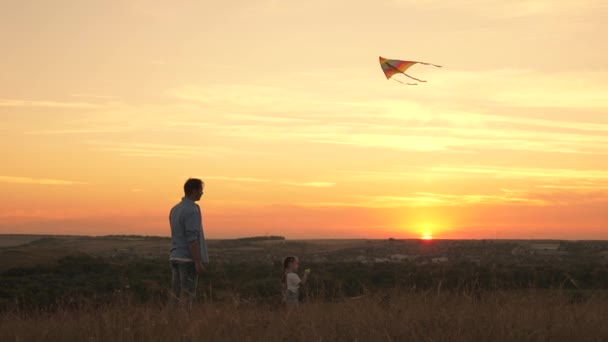 幼い娘と一緒に公園の日没時に凧で遊んでいる父親。屋外ファミリーゲーム。父と健康な娘は多色の紙飛行機を空に打ち上げます。子供は凧を飛ばしてる。幸せな家族 — ストック動画