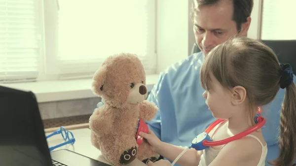 Cuidar a un pediatra profesional masculino jugando con un niño pequeño en la oficina. niña juega con peluche juguete le escucha con un estetoscopio. niño pequeño paciente se sienta en el regazo de pediatra. —  Fotos de Stock