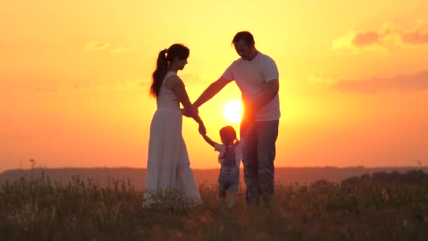 Joven mamá, papá, con una pequeña hija sana están bailando en un círculo en el sol caliente, divirtiéndose en el campo. familia feliz tomados de la mano juega en el parque en la hierba, al atardecer. Concepto de infancia e infancia — Vídeos de Stock