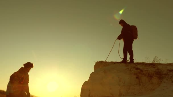 Scalatori scalano la montagna su una corda. un turista maschio aiuta una ragazza viaggiatore a scalare la montagna. Viaggio e avventura in montagna al tramonto. lavoro di squadra di turisti al sole. concetto di libertà e avventura — Video Stock