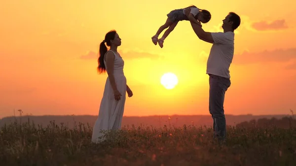 Papa speelt met zijn dochtertje, gelukkig om kind in de lucht te gooien, gelukkige moeder bewondert haar familie. Mama papa en baby, familie rust uit bij zonsondergang in het veld. gelukkig gezond gezin wandelen in de frisse lucht. — Stockfoto