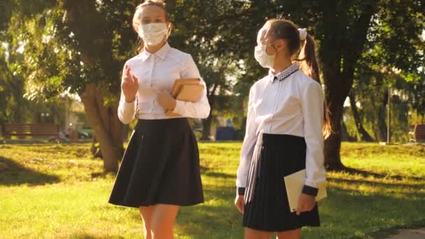 A lányok az utcán sétálnak maszkban, tankönyveket és tabletet tartanak a kezükben. A tinédzserek a parkban járnak iskolába. lányok sétál orvosi maszkok elleni védelem vírusok és baktériumok. — Stock videók