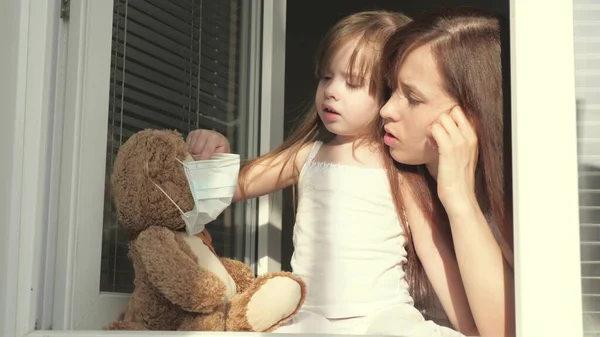 Mom and daughter play on windowsill, put on a protective mask from viruses on teddy bear, play and look out open window. childs family and mother are in home quarantine. Covid-19 coronavirus — Stock Photo, Image