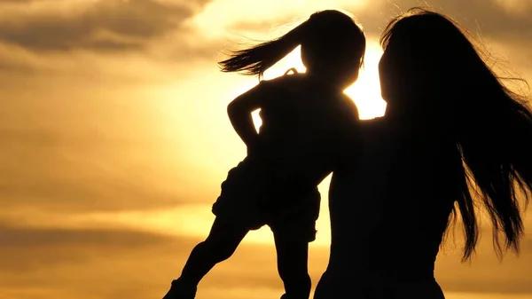 Mamá está rodeando a su querida hijita sana en sus brazos. silueta de mamá y el bebé al atardecer. madre feliz y el niño pequeño abrazándose en el parque en la naturaleza en los rayos del sol. familia de vacaciones. —  Fotos de Stock