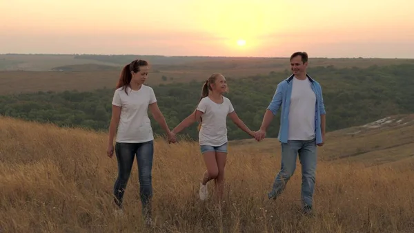 Hälsosam leende familj som håller händer gå i fält vid solnedgången i bergen, dotter håller mamma och pappa i händerna. glada barn och föräldrar gå i strålar av vacker sol, resa på semester — Stockfoto