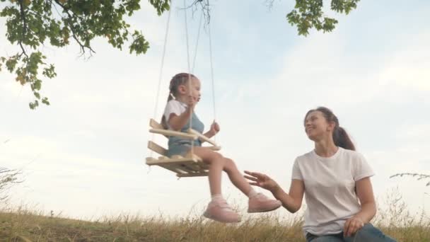 La familia libre se está divirtiendo en el parque. madre sacude a su pequeña hija sana en un columpio bajo un árbol en el sol. madre juega con su hijo, se balancean en la cuerda en una rama de roble en el bosque. — Vídeos de Stock