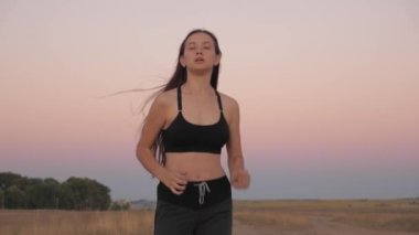 Sağlıklı, güzel bir kız spor yapıyor, kırsalda güneş altında koşuyor. Hayat devam ediyor. Antrenman koşusu. Koşucu kız sahada temiz hava soluyor. Özgür genç kadın gün batımında yaz parkında koşuyor..