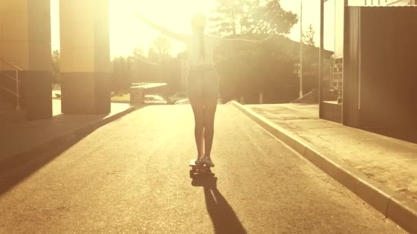 Беззаботный подросток катается на доске в городе с протянутыми руками. Девушка учится кататься на скейтборде на улице под солнцем. Счастливый скейтбордист едет по дороге в город на закате. — стоковое видео