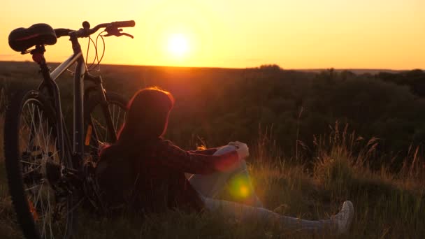 Gezonde jonge vrouw wandelaar zittend op de heuvel naast de fiets, genietend van natuur en zon. concept van avontuur en reizen. Alleenstaande fietser rustend in het park. gratis meisje reist met een fiets in zonsondergang. — Stockvideo