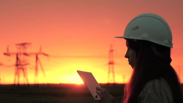 Seorang insinyur listrik wanita dalam helm putih memeriksa kabel listrik menggunakan data dari sensor listrik pada tablet. Tegangan tinggi saluran listrik saat matahari terbenam. Distribusi dan pasokan listrik. bersih energi — Stok Video