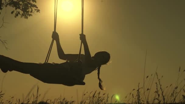 Silhouette of a small healthy child on a swing. child swinging on a swing in park in sun. teen girl enjoys flight on swing on summer evening in forest. concept of happy family and childhood. — Stock Video