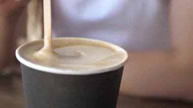 Masanın üzerinde bir bardak kahve var, bir kız lezzetli aromalı kahve karıştırıyor. Yakın plan. Kahve, sıcak bir içecek için tahta kaşığı karıştıran adam..