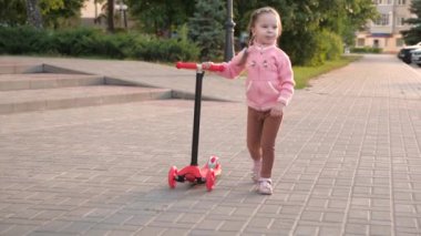 Sağlıklı bir çocuk sokakta scooter kullanıyor. Mutlu çocukluk kavramı. Küçük kız scooter sürmeyi öğreniyor. Parkta oynayan mutlu kız bebek.