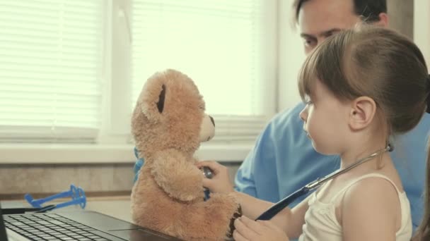 Klein meisje speelt met een teddybeer speelgoed luistert naar hem met een stethoscoop. peuter patiënt zit op schoot van kinderarts. Zorgvuldige professionele mannelijke kinderarts spelen met een klein kind in het kantoor. — Stockvideo