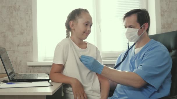 Médico masculino pone un estetoscopio en el pecho de un niño, sonríe y escucha a una niña. pediatra médico examina niño. El médico escucha los latidos del corazón del bebé. Un niño en una habitación de hospital. — Vídeo de stock