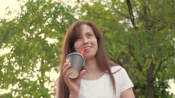 Mujer feliz bebiendo café aromático caliente al aire libre, riendo, hablando y sonriendo. Joven mujer de negocios hermosa está caminando en el parque de la ciudad con una taza de delicioso té para llevar. — Vídeo de stock