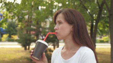 Genç ve güzel bir iş kadını, şehir parkında bir fincan lezzetli kahve eşliğinde gülümsüyor. Mutlu kız dışarıda sıcak aromatik çay içiyor..