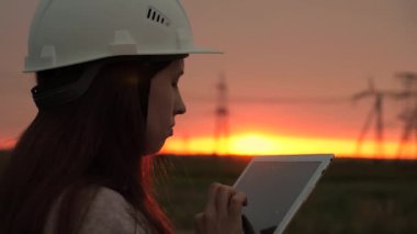 Kadın mühendis güç mühendisi. Gün batımında yüksek voltajlı elektrik hatları. Temiz enerji. Beyaz kasklı genç uzman kız tabletteki elektrik sensörlerinin verilerini kullanarak güç hattını kontrol ediyor..