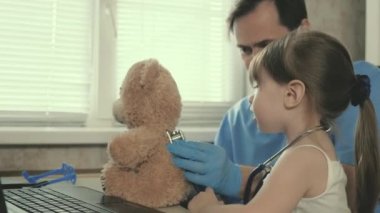 Yeni yürümeye başlayan hasta çocuk doktorunun kucağına oturuyor. Ofiste küçük bir çocukla oynayan profesyonel bir erkek çocuk doktoru. Küçük kız oyuncak ayıyla oynuyor ve steteskopla onu dinliyor..