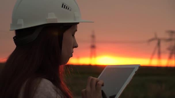 Femme ingénieur inspecteur ingénieur de puissance. Lignes électriques à haute tension au coucher du soleil. énergie propre. jeune fille spécialisée dans le casque blanc vérifie la ligne électrique à l'aide de données de capteurs électriques sur tablette. — Video