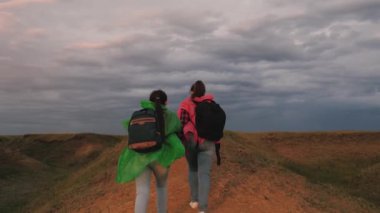Özgür kadın gezginler tepeye tırmanıyor, ellerini kaldırıyor ve zaferlerine seviniyorlar. Gezginlerin takım çalışması. Macera ve seyahat. Sağlıklı turistler sırt çantaları ve renkli yağmurluklarla seyahat ederler..