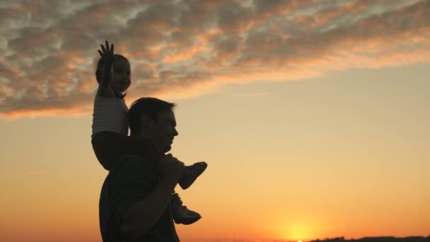 Pappa bär på sina älskade barns axlar, solstrålar. barn med föräldrar går vid solnedgången. Pappa går med sin dotter på axlarna i solnedgångens strålar. lycklig familj vilar i parken. — Stockvideo